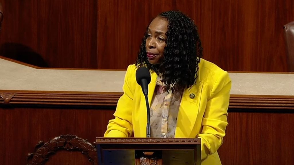 Yvette Clarke in a yellow blazer speaks at a podium in a formal setting.