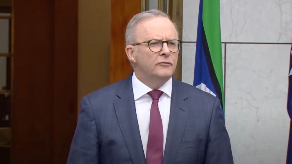 Anthony Albanese in a suit and tie standing in front of flags, speaking outdoors.