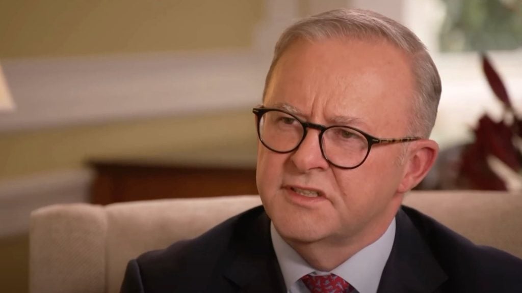 Albanese wearing glasses and a suit is sitting on a couch, appearing to engage in a conversation.