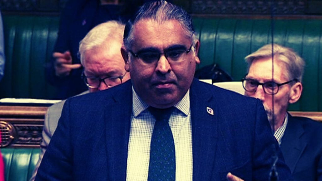 MP ALI wearing a suit and glasses is speaking in a formal setting, with other individuals seated in the background.