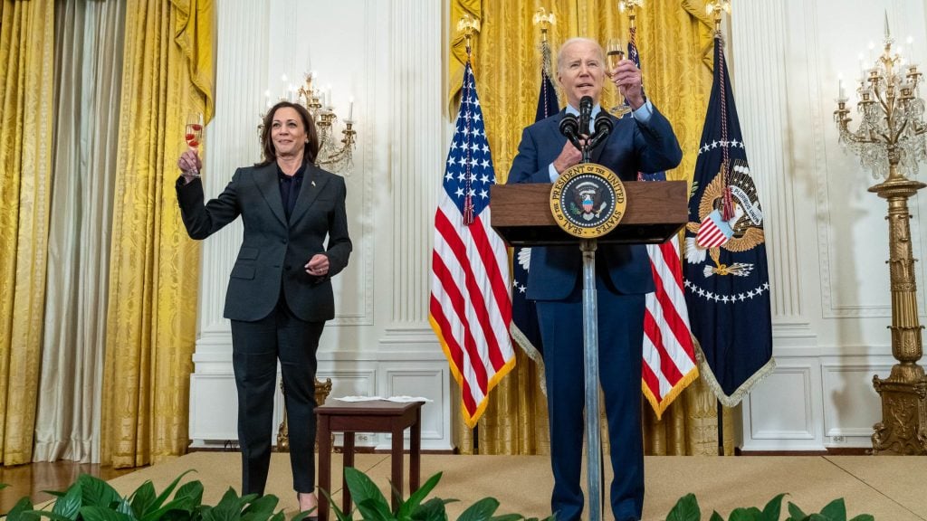 Biden and Harris in a room with gold curtains, holding toasting glasses, with U.S. flags and a presidential podium.