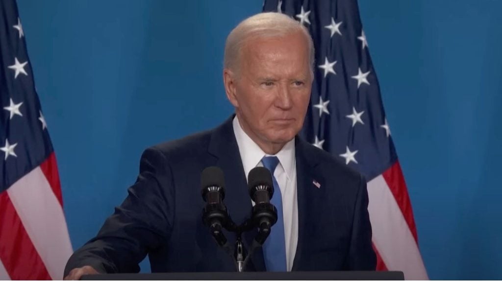 Biden in a suit stands at a podium with microphones, with USA flags in the background.