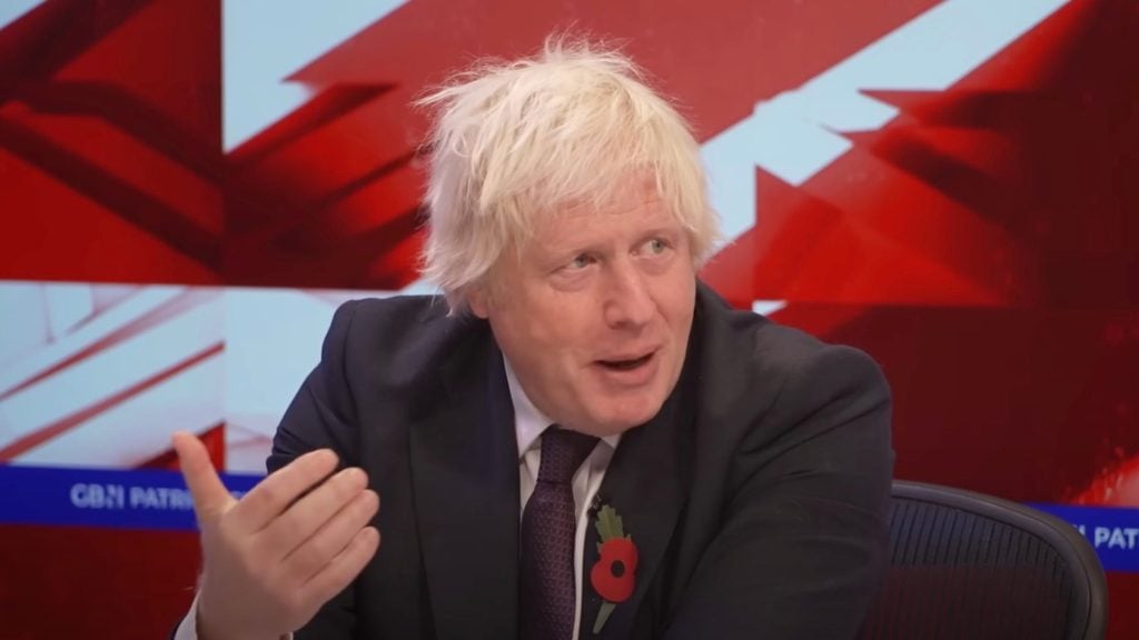 Boris Johnson in a suit and tie talking, wearing a red poppy lapel pin, in front of a red and white background.