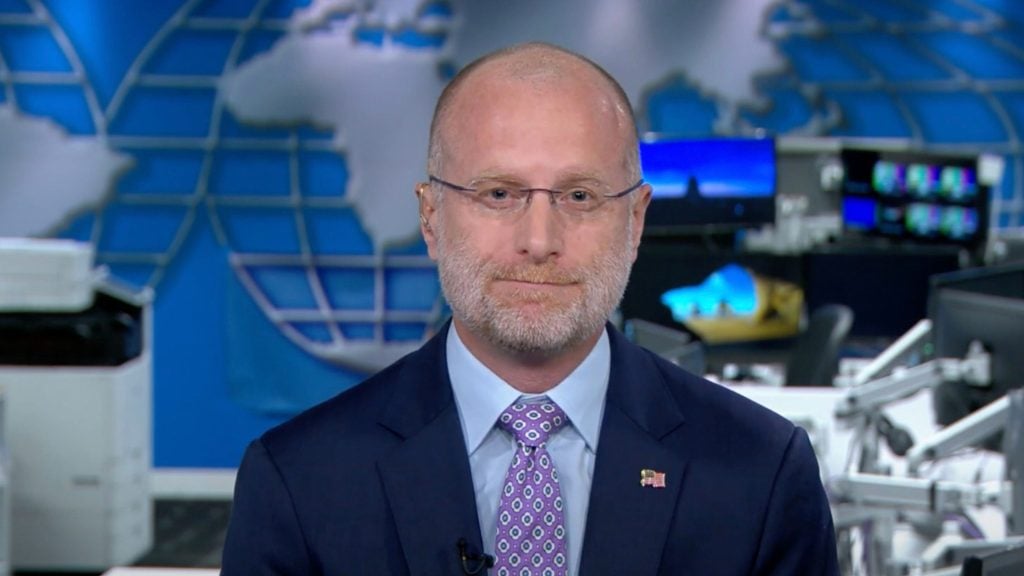 Carr with a beard wearing a suit and patterned tie, standing in a news studio.