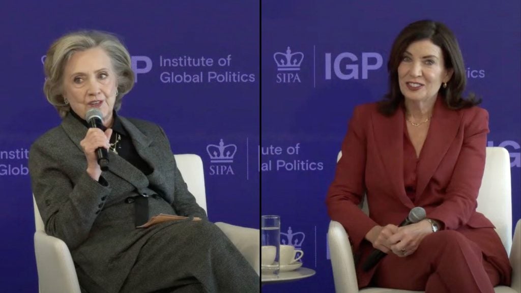 Hillary Clinton, Kathy Hochul seated on stage at the Institute of Global Politics event, both holding microphones, one wearing a gray suit and the other a red suit.