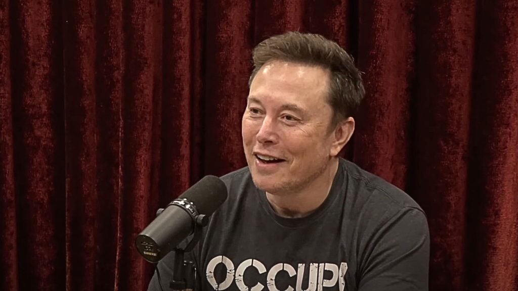 Elon Musk speaking into a microphone with a red curtain background.