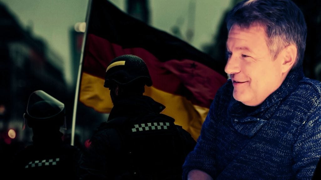 Habeck smiling with a blurred background featuring a person in riot gear and a German flag.