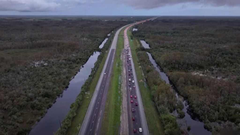 Aerial view of a long highway surrounded by dense forest and waterways, with a heavy traffic jam on one side.