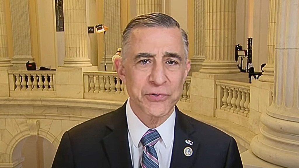 Issa in a suit standing in a grand interior with marble columns and ornate railings in the background.