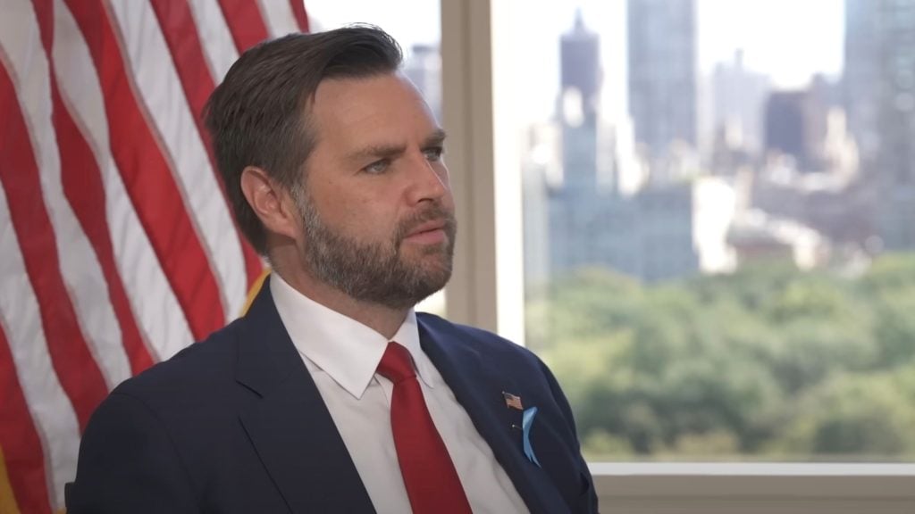 JD Vance in a suit with an American flag pin, sitting in front of a US flag, blurred cityscape in the background.