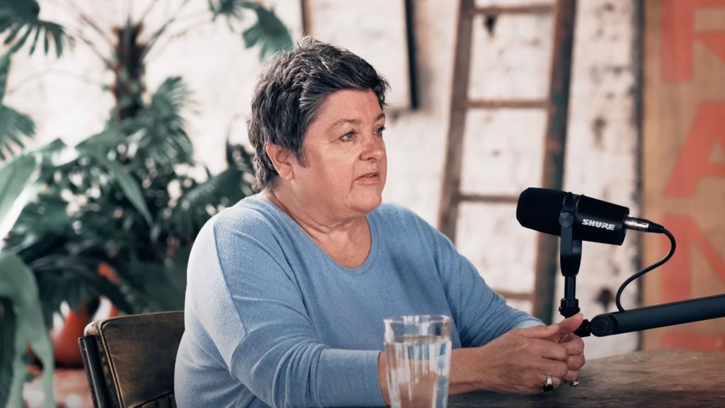 Julie Bindel in a blue shirt sits at a table with a microphone, speaking and gesturing with her hands.