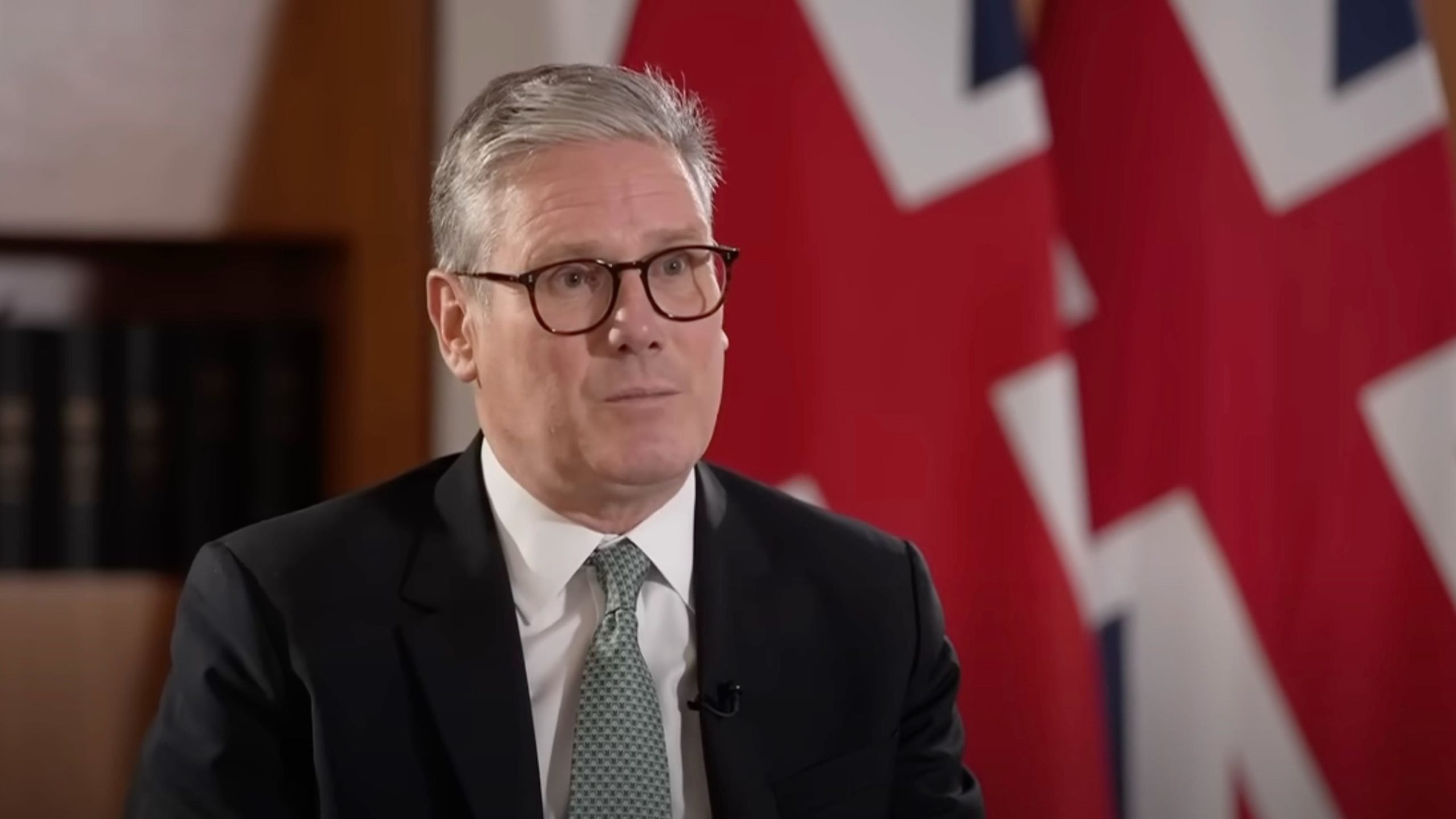 Keir Starmer in a suit and glasses speaking, with a British flag in the background.