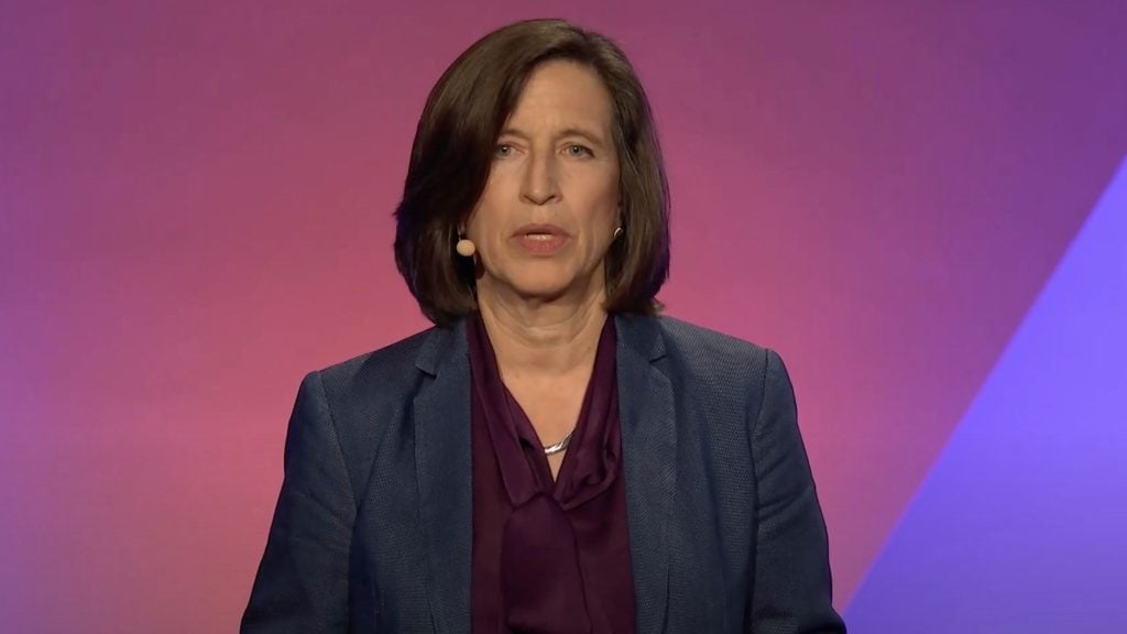 Melissa Fleming with shoulder-length brown hair wearing a blue blazer and purple blouse, with a headset microphone, against a pink and purple background.