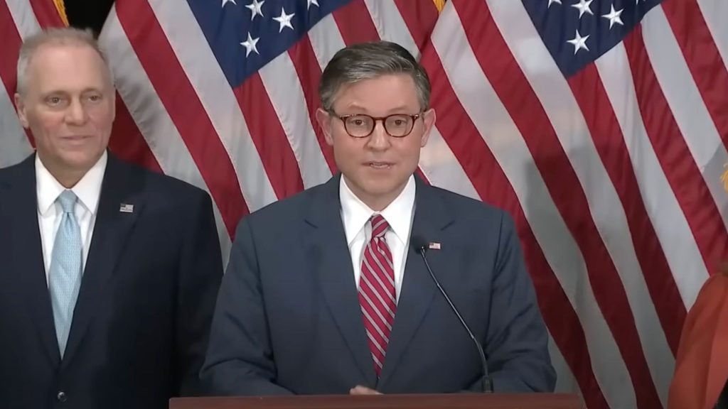 Mike Johnson in a suit stands in front of American flags, with one speaking at a microphone.