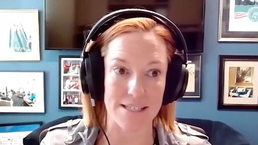 Psaki with headphones speaking in front of a wall with framed pictures.