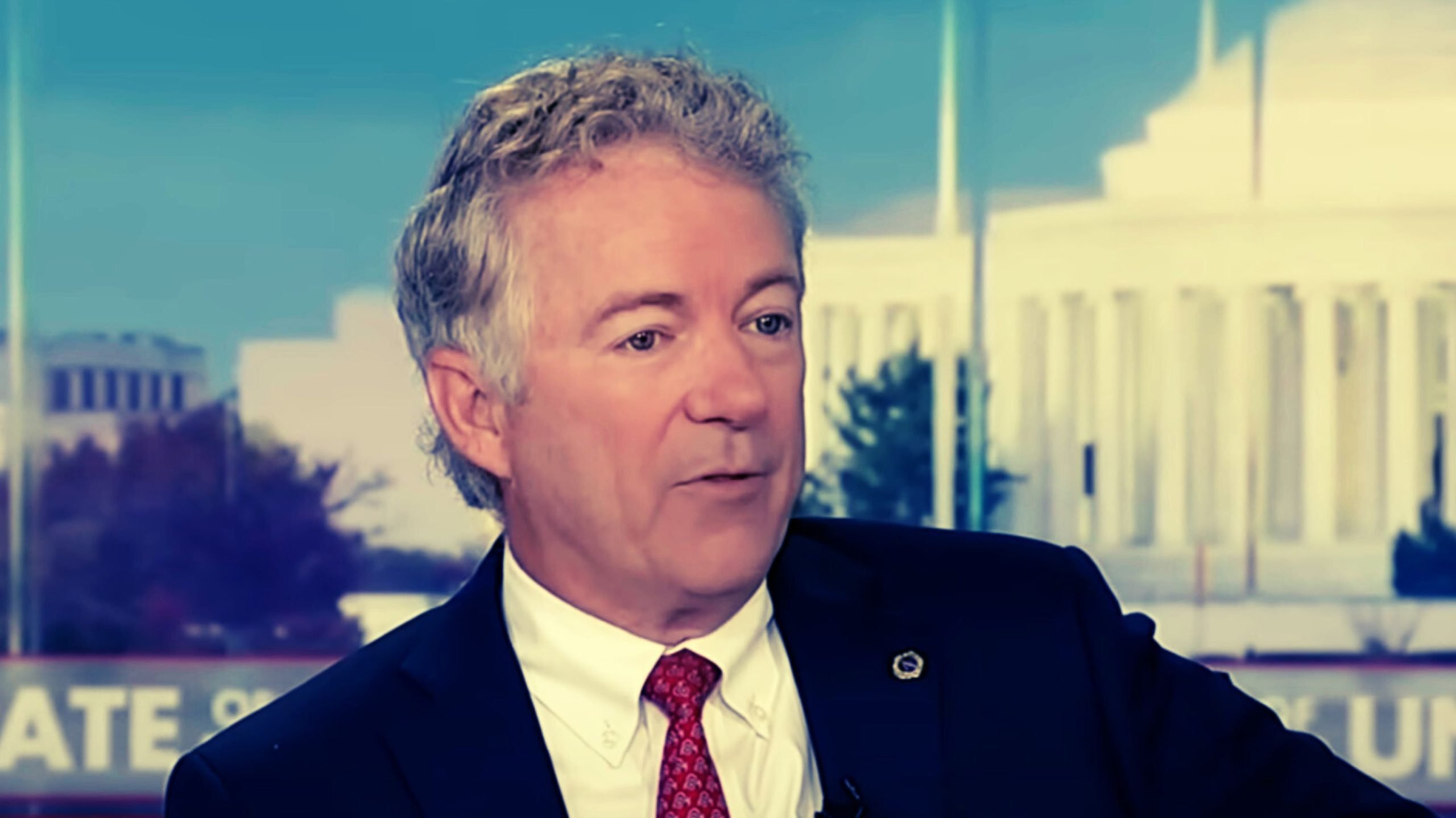 Rand Paul in a suit and red tie appears to be speaking on a news program, with a blurred cityscape in the background.