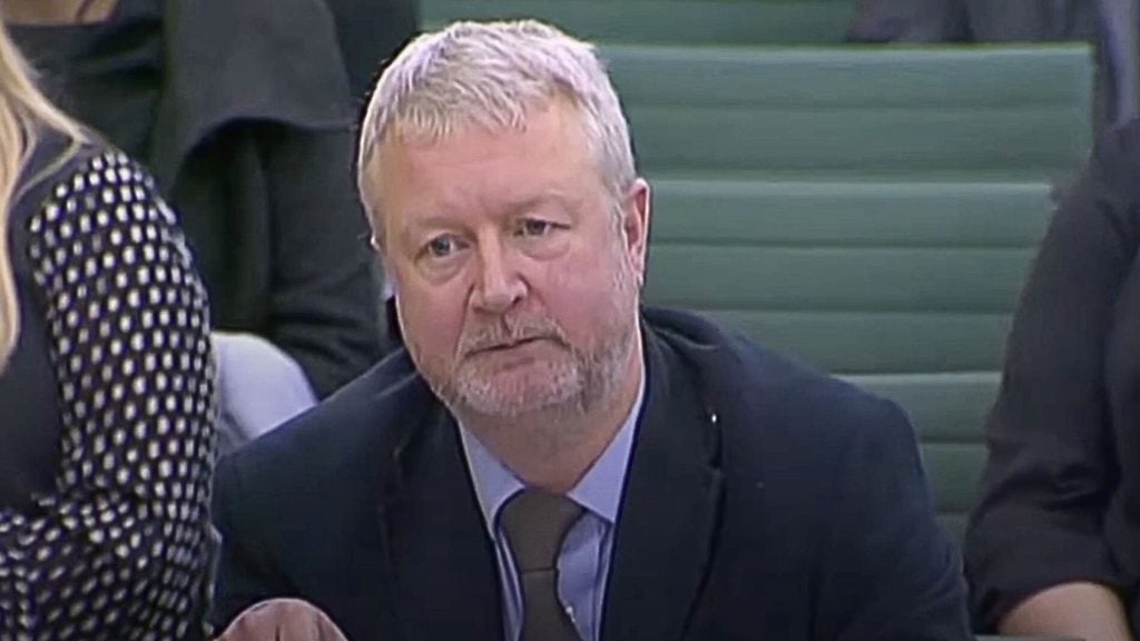 Lord Richard Allen with short gray hair and a beard, wearing a suit and tie, seated indoors.