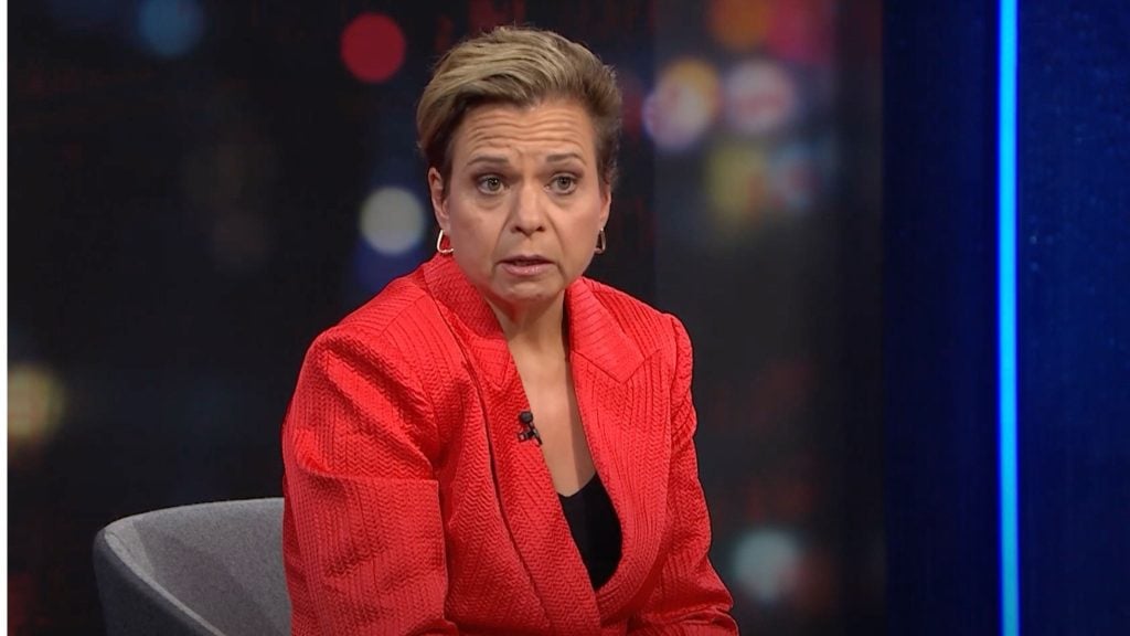 A person seated in a chair, wearing a red blazer, with a blurred background.