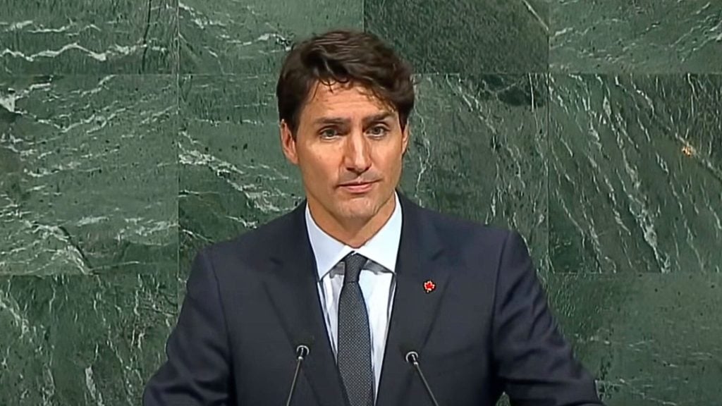 Justin Trudeau in a suit speaking at a podium with a green marble background.