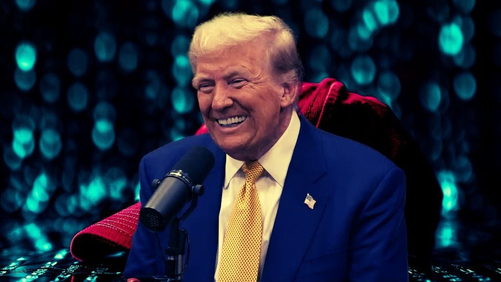 President Trump in a blue suit with a yellow tie sits in front of a microphone, smiling, with a blurred, colorful background.