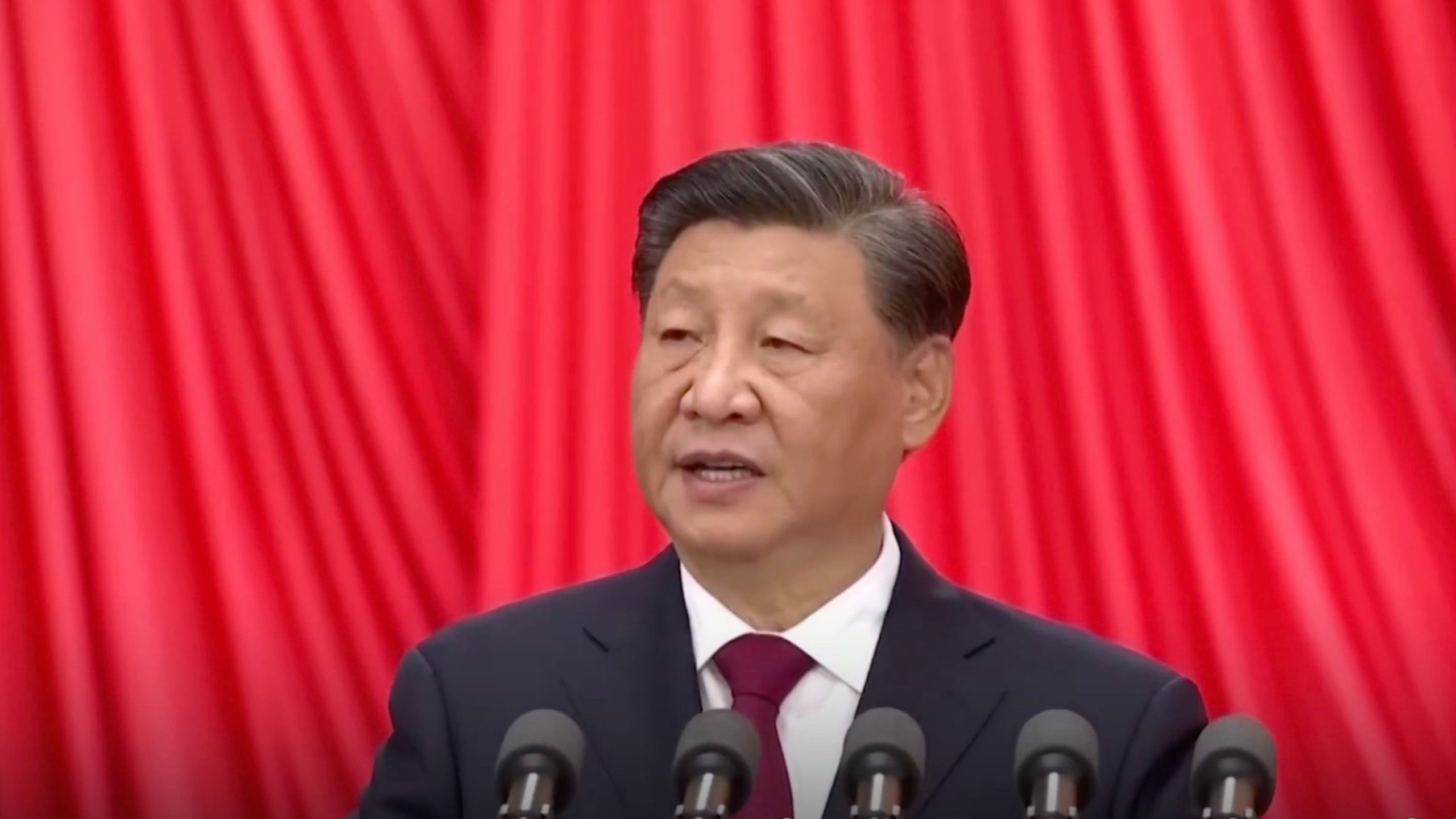 Xi Jinping in a suit speaking at a podium with a red curtain background.