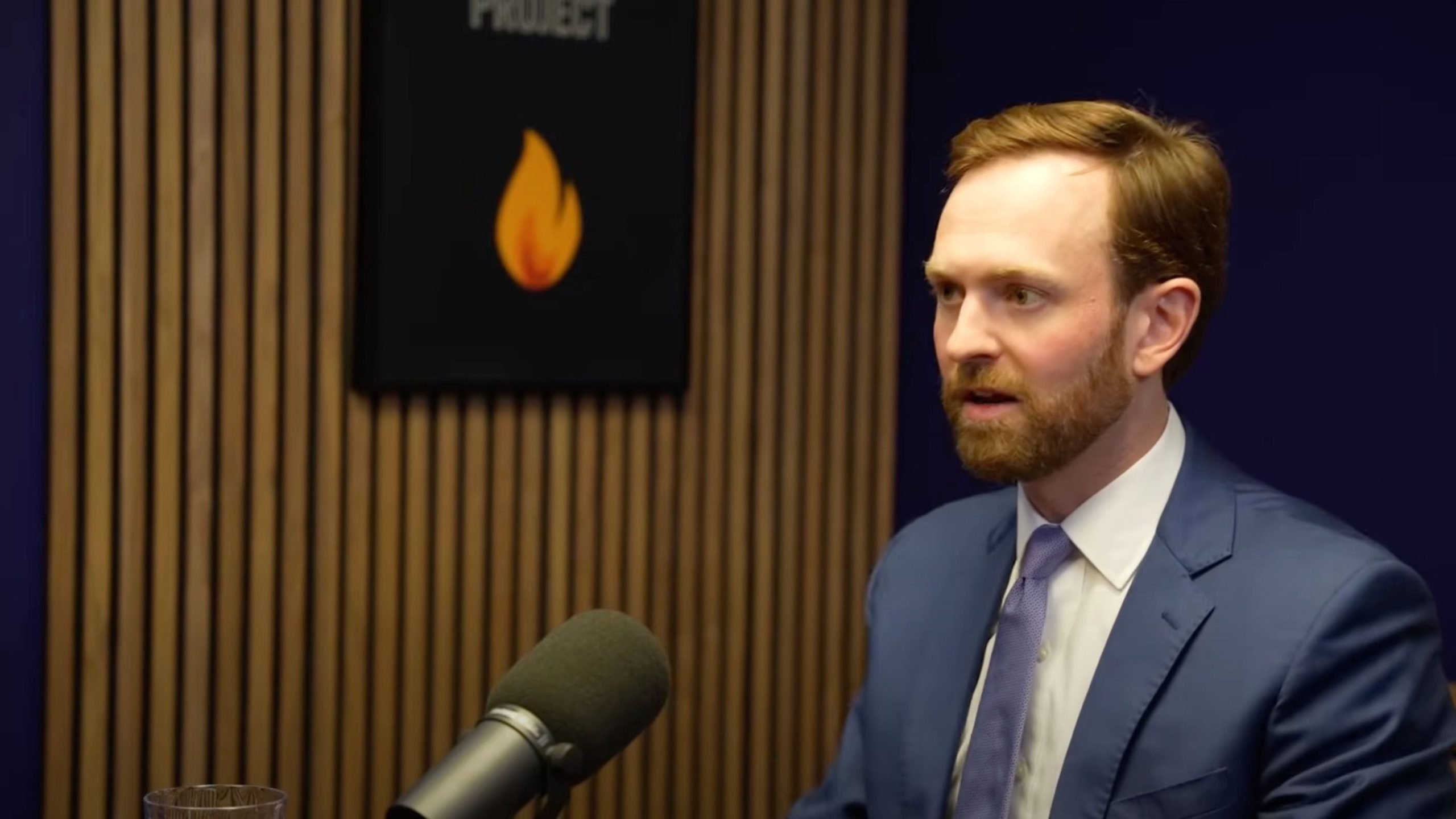 Ferguson in a blue suit speaking into a microphone in a podcast studio with wooden panels.