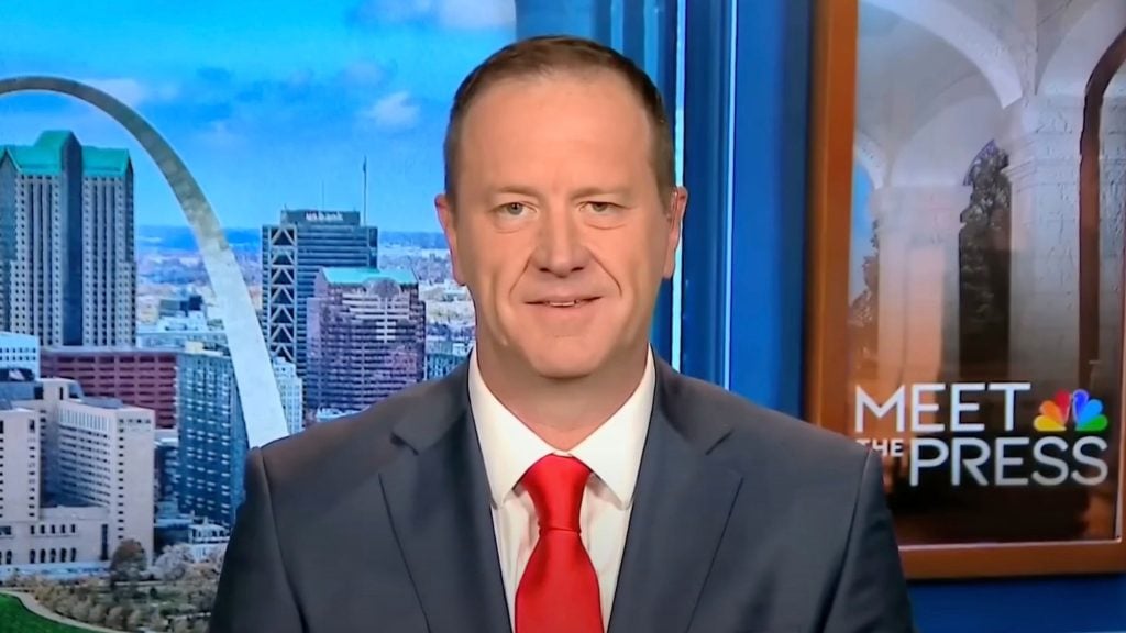 Eric Schmitt in a suit and red tie is on a TV program with a cityscape and the Gateway Arch in the background. The show's logo, "Meet the Press," is visible.