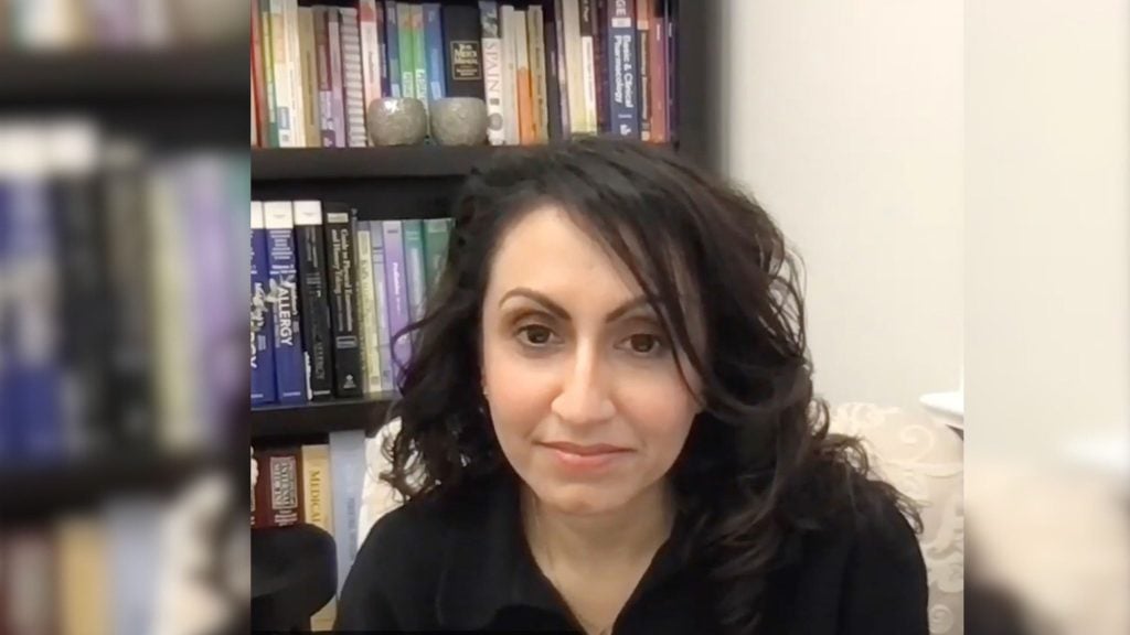 Kulvinder-Kaur-Gill with dark hair sits in front of a bookshelf filled with various books.