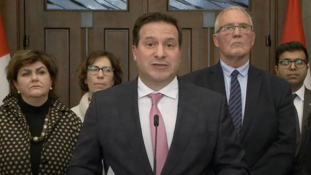 Marco Mendicino standing at a podium, with a man in a suit and pink tie speaking into a microphone.