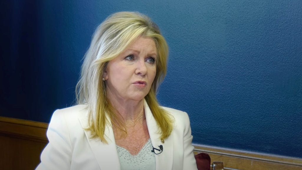 Blackburn with blonde hair wearing a white blazer sits in front of a blue background.