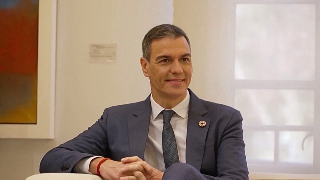 Sanchez seated indoors, smiling with hands clasped, wearing a red bracelet and a pin.