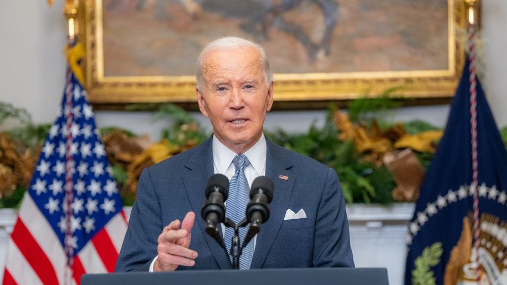 Biden in a suit speaking at a podium with two microphones, American flag and executive symbol in the background.