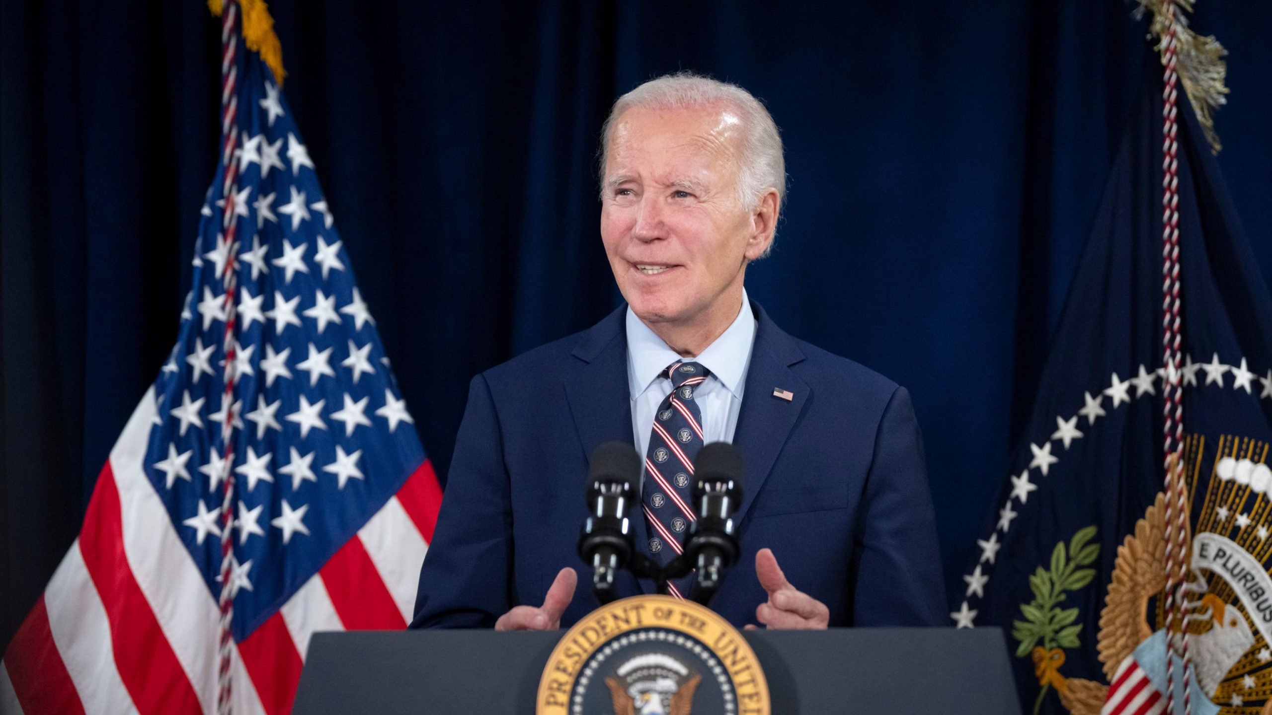 Biden in a suit speaking at a podium with American flags in the background.