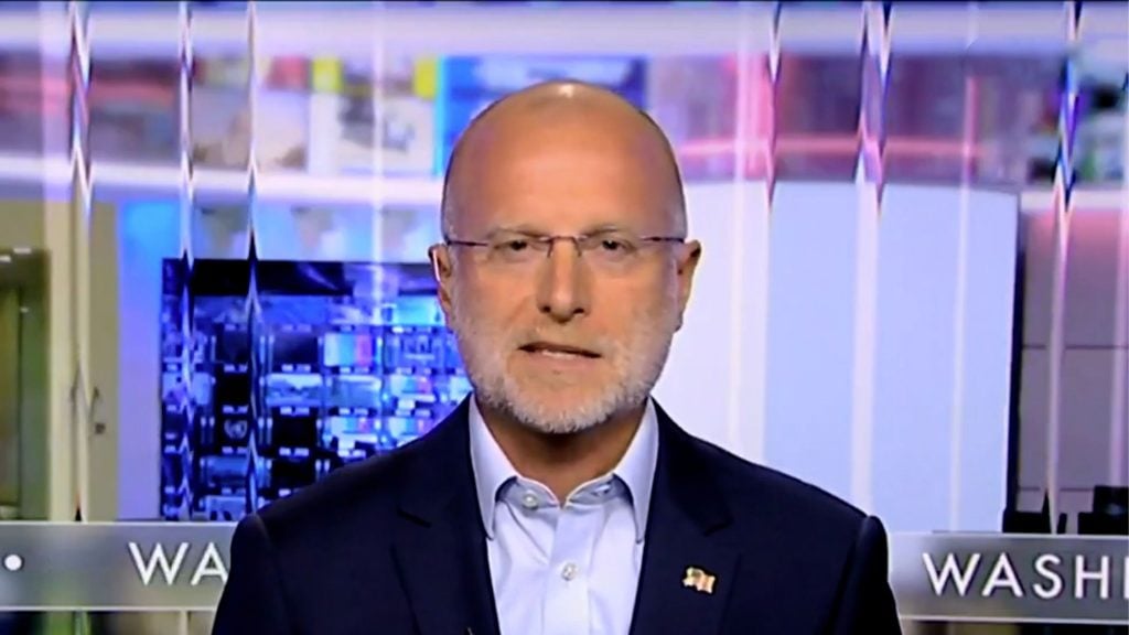 Brendan Carr in glasses speaking on a television news segment, with a blurred background and the text "WASHINGTON" partially visible.