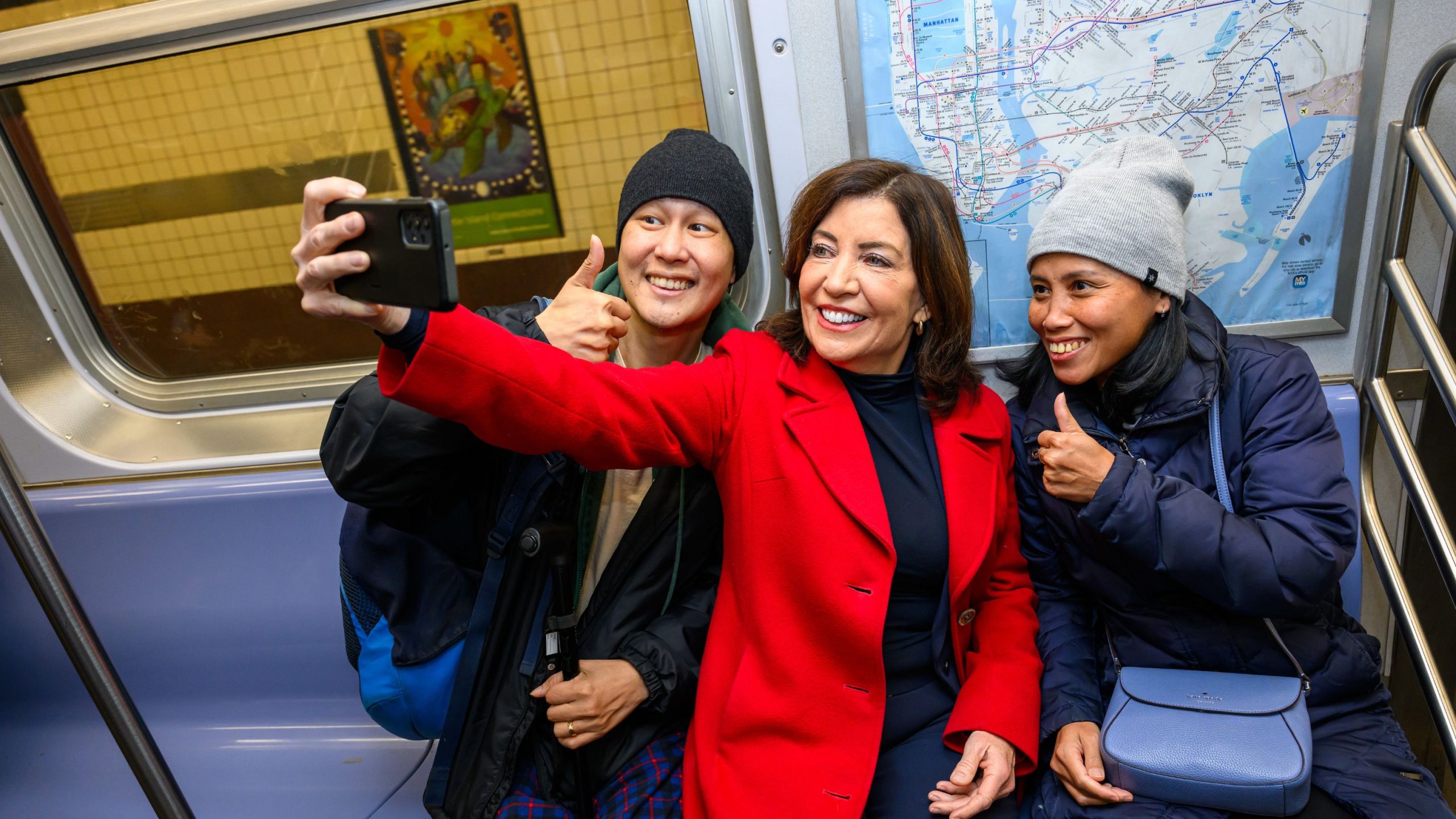 Hochul taking a selfie while sitting in a subway car, with one person in a red coat holding the phone.