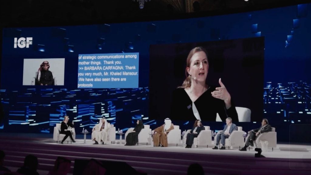 A panel discussion on stage with multiple speakers seated, large projected screens showing a close-up of a woman speaking, and text displaying "IGF".