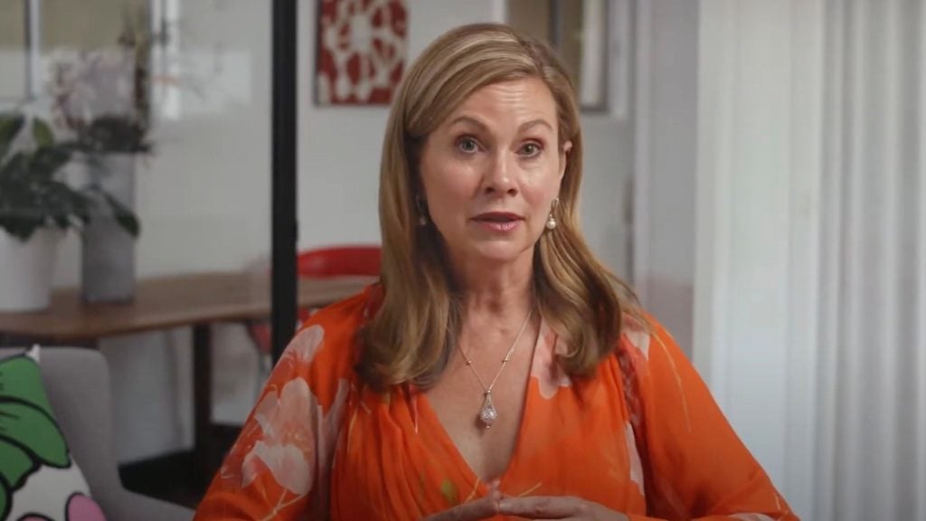 Julie Inman Grant with long blonde hair wearing an orange top and a necklace, seated indoors with a blurred background.