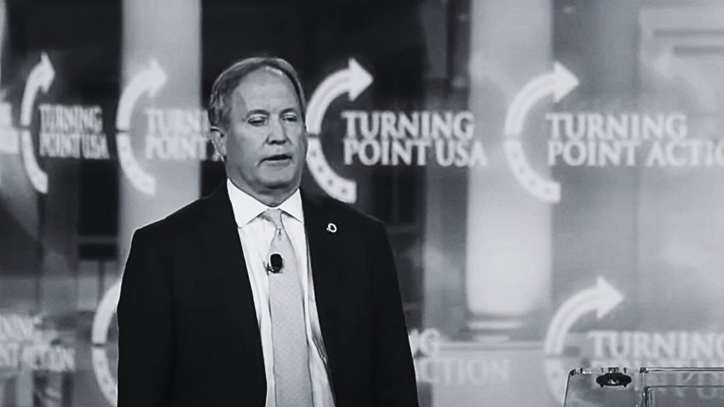 Paxton in a suit and tie stands in front of a Turning Point USA backdrop, speaking at a podium.