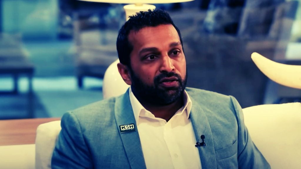 Kash Patel in a blue suit with a lapel pin, sitting indoors with a blurred background.