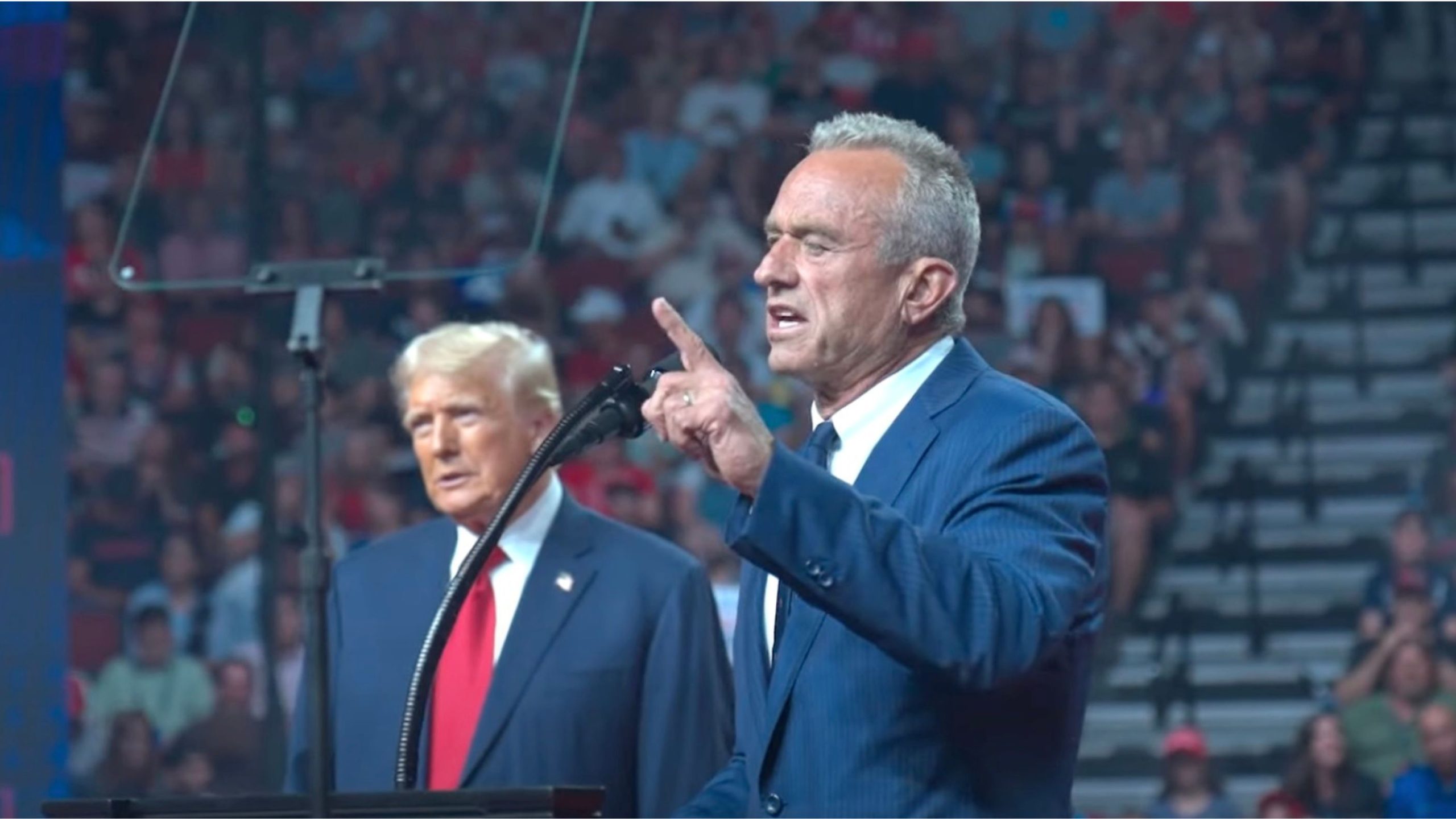 Trump and Kennedy in suits speaking at a large indoor event with an audience.