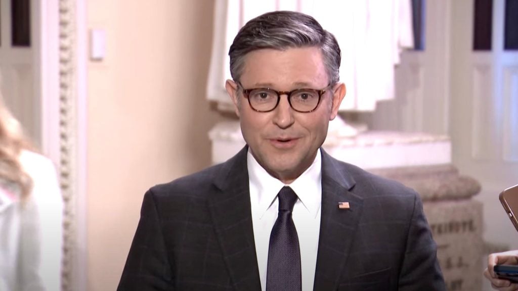 Mike Johnson in a suit, and a tie with an American flag pin, speaking indoors.