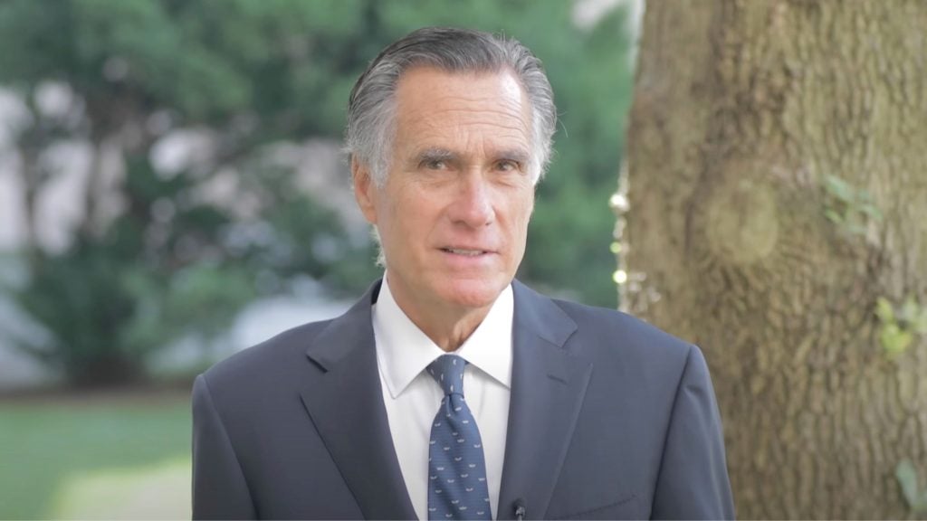 Romney in a suit standing outdoors near a tree with greenery in the background.