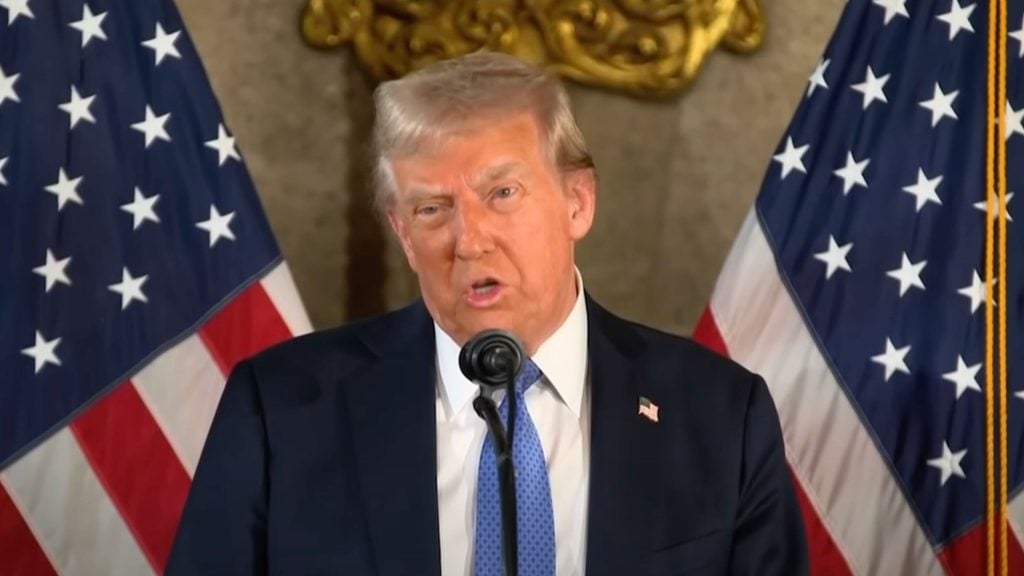 Trump speaking at a podium with two American flags in the background.