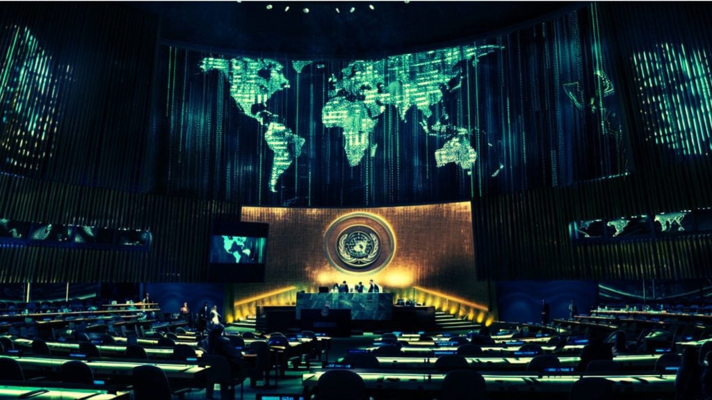 A large conference room with a digital world map displayed on screens, featuring a circular emblem on the wall behind a panel of speakers at a podium, and rows of seating below.