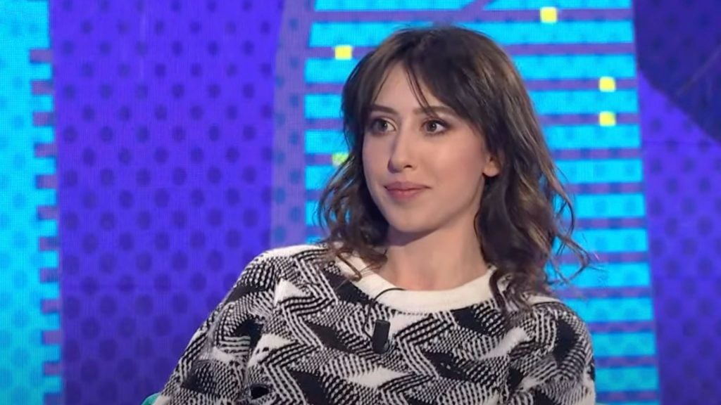 Cecilia Sala with shoulder-length brown hair wearing a black and white patterned sweater is sitting in front of a colorful, geometric background.