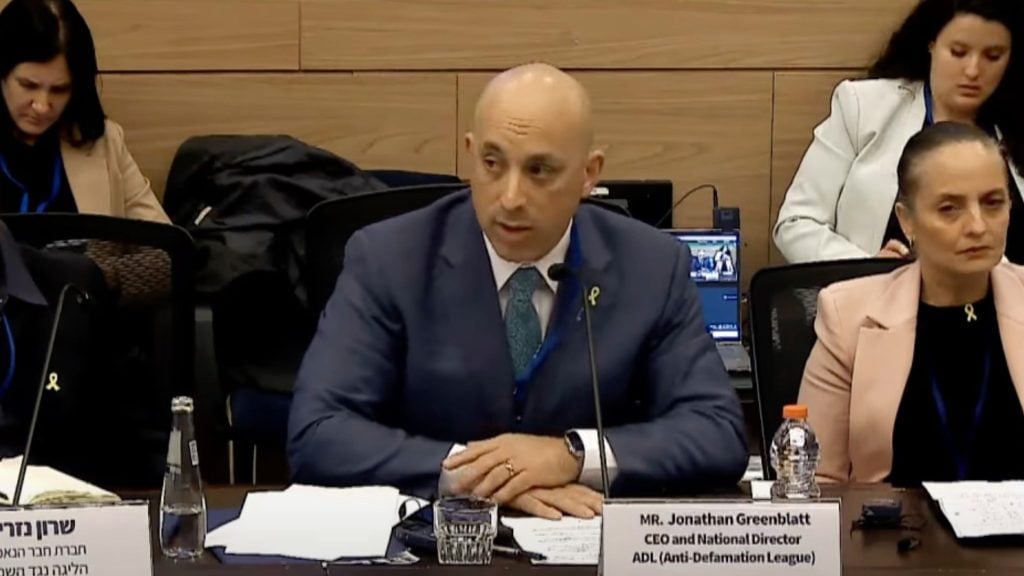 Greenblatt in a suit is speaking at a conference table with a microphone; nameplate in front reads "Mr. Jonathan Greenblatt." Two women are seated nearby.