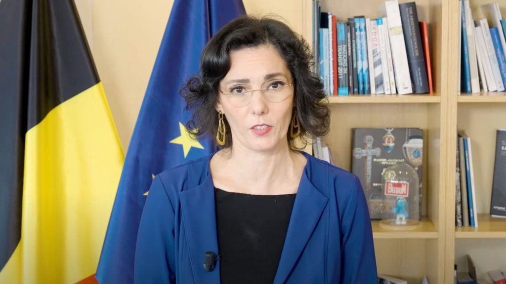 Hadja Lahbib in a blue jacket stands in front of EU and Belgian flags, with a bookshelf in the background.