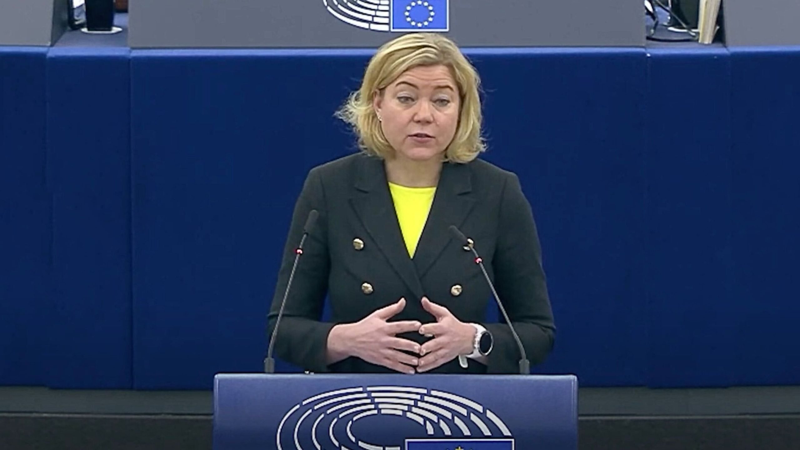 Henna Virkkunen in a black double-breasted blazer speaks at a podium with European Union symbols.