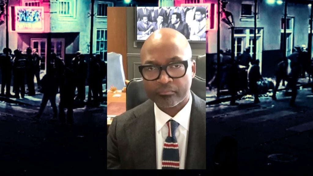 Jason Williams wearing glasses and a suit is sitting in an office, with a black and white historical photo on the wall behind them.