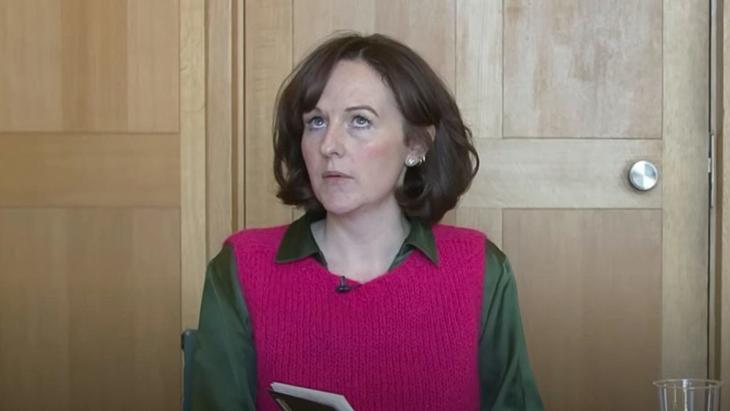 McEvoy with short brown hair wearing a pink sweater vest over a green shirt is looking upward.